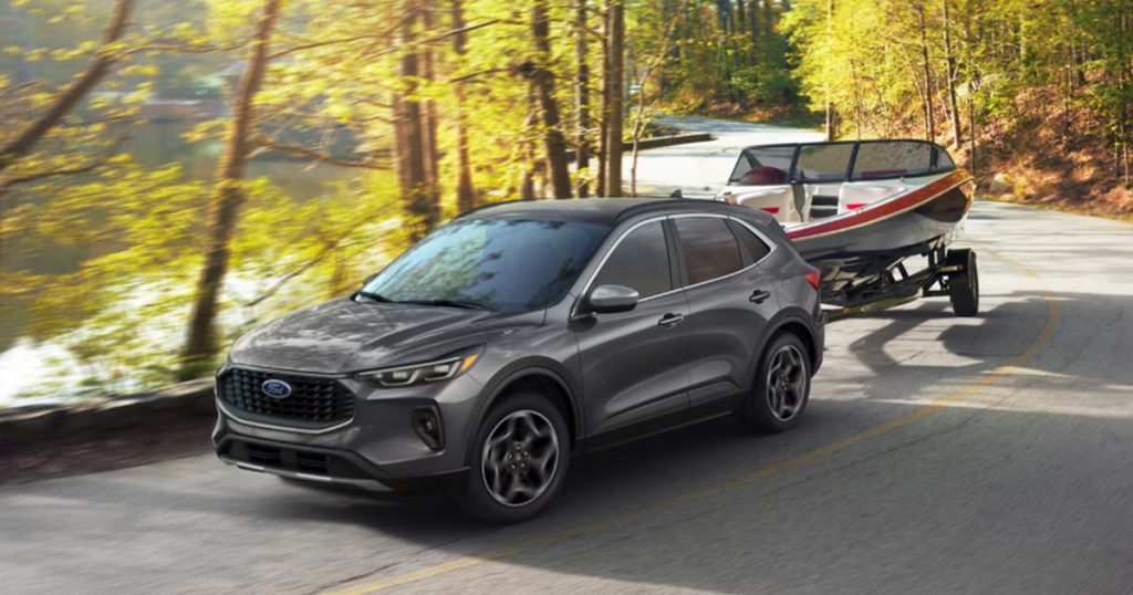 A dark grey 2026 Ford Escape travels down a tree-covered road towing a boat behind it.